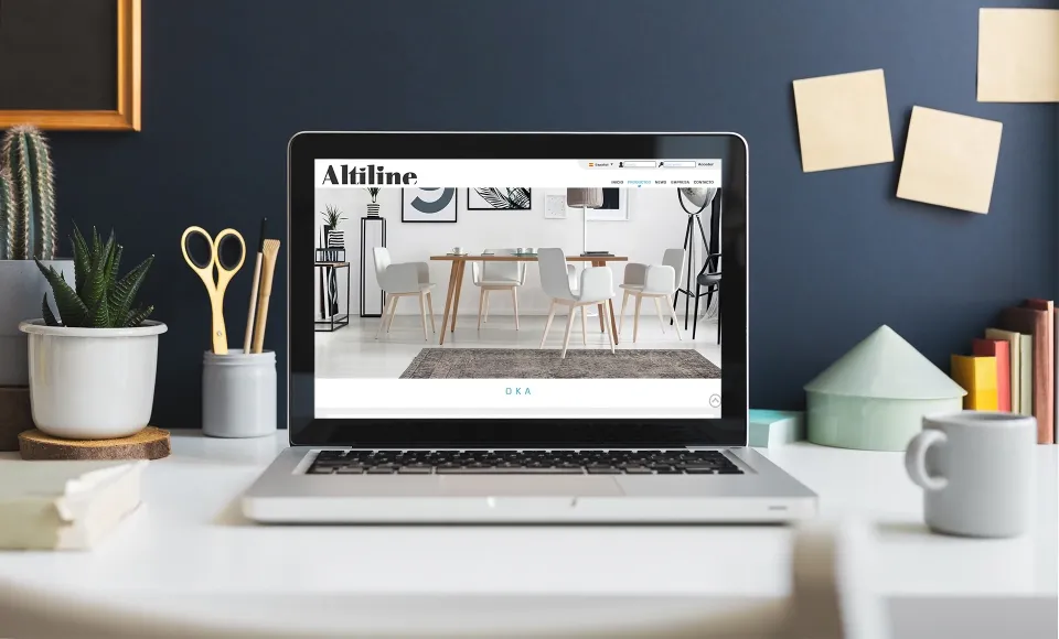 Web de Altiline en un portátil sobre un escritorio, con un comedor de mesa y sillas de madera